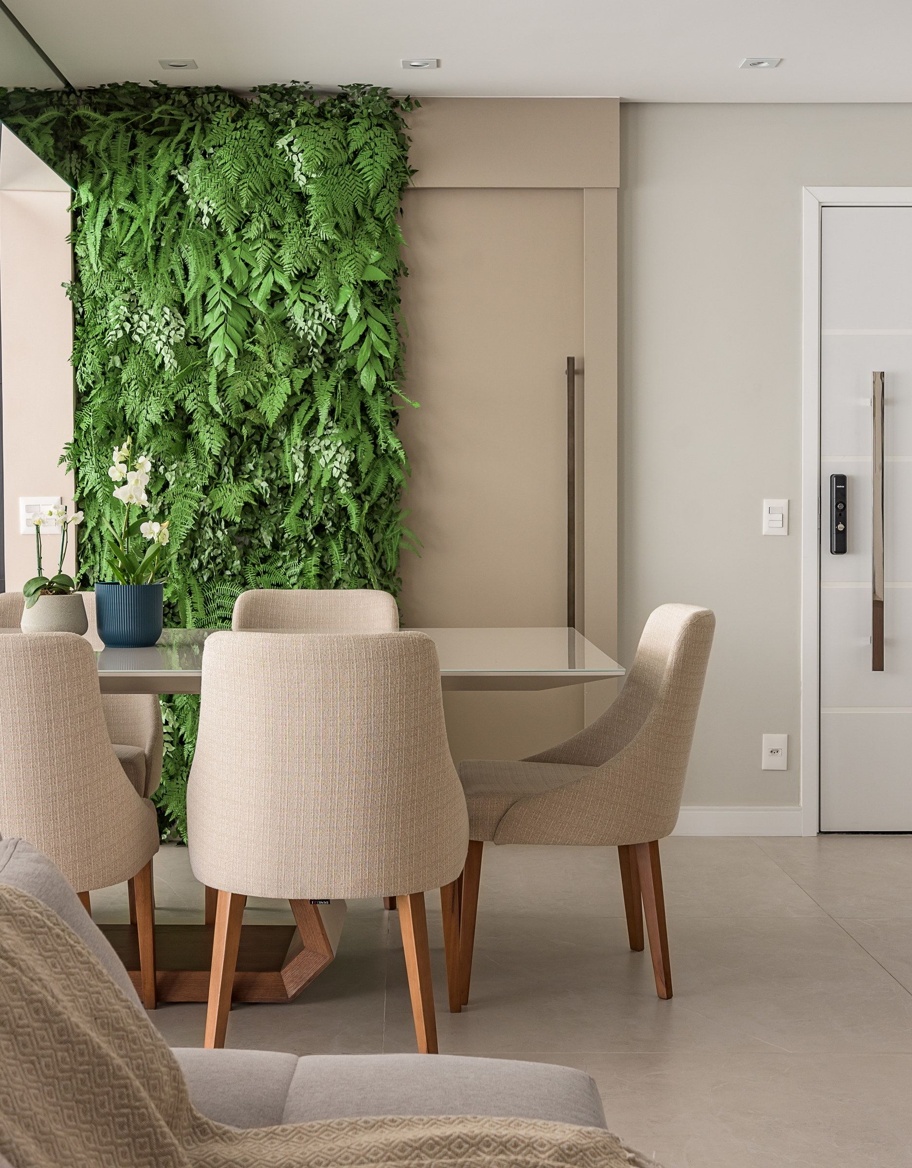 Sala de jantar com jardim vertical de samambaias na parede, mesa de vidro e cadeiras bege