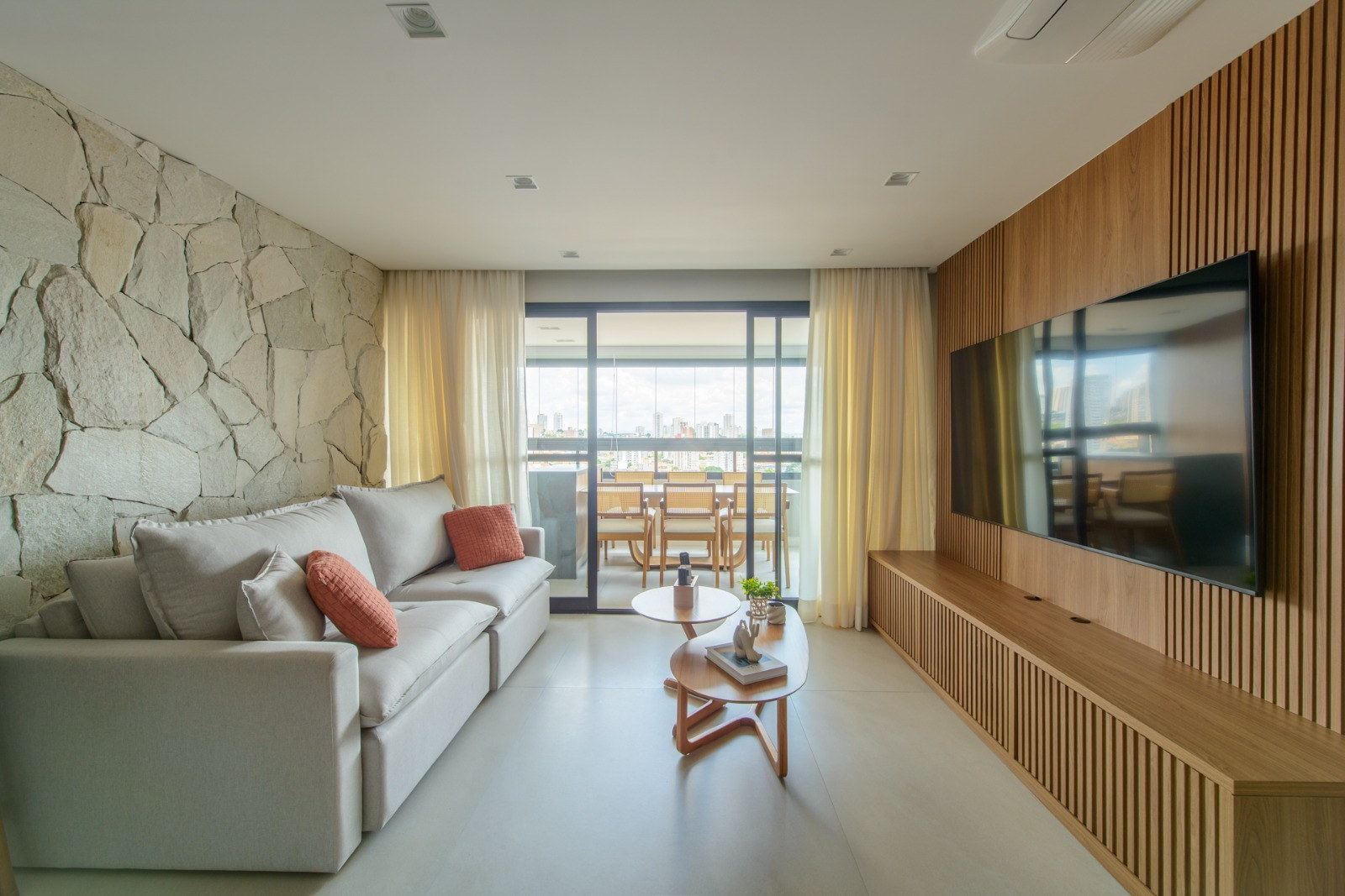 Sala de estar entre parede de pedra natural e painel ripado de madeira, com varanda envidraçada ao fundo